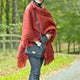 A person stands in a wooded area, wearing a red shawl, black pants, and gloves, holding a quilted black purse on a path surrounded by greenery.