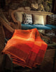 Yacupungu baby alpaca wool blanket – a vibrant orange-striped blanket drapes over the arm of a wicker chair. In the background, a cozy couch with assorted cushions sits against a stone wall