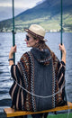 A person wearing a patterned poncho and hat sits on a swing above water, with a mountainous landscape in the background.