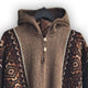 A brown knitted hooded sweater with two buttons, featuring intricate geometric patterns on the sleeves, is hanging on a black hanger against a white background.