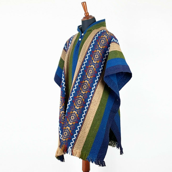 A multicolored poncho with geometric patterns hangs on a mannequin, featuring blue, green, beige stripes and fringed edges, set against a plain white background.