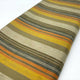 Rumi baby alpaca wool blanket – fabric with horizontal stripes in earthy tones of yellow, orange, brown, and beige, is laid out flat. The texture appears woven, and the fabric extends across a plain background