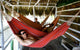 Three people relax in colorful hammocks under a thatched roof hut on a wooden deck, surrounded by wooden railings and beams, suggesting a tropical, leisurely setting.
