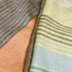 Latacunga alpaca wool throw blanket – fabrics drape smoothly, featuring soft pastel hues with alternating horizontal stripes in brown, yellow, and blue tones. The background is textured with a peach-colored surface