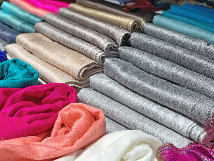Rolls of colorful fabric, including shades of gray, beige, and vibrant pinks, are neatly stacked. Surrounding silk and woolen textures suggest a textile market or fabric store setting.