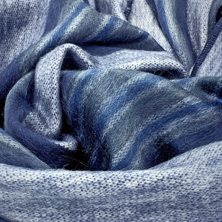 Chimborazo baby alpaca wool blanket – a textured fabric displays intertwined folds in shades of blue and grey, forming abstract patterns. The woven material is shown in close-up, highlighting its soft, fibrous texture