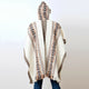 A person stands wearing a hooded poncho, featuring geometric patterns in brown and black, with frayed edges. The background is a plain, light-colored wall.