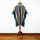 A poncho with blue, green, and beige stripes hangs on a wooden stand; intricate geometric patterns adorn it, set against a white wall and wooden floor.