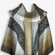 A woolen poncho with intricate geometric patterns in shades of brown and gray is draped symmetrically. The fringed edges add texture, while a plain background enhances its detail.