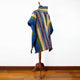 A colorful woven poncho is displayed on a wooden mannequin stand, featuring geometric patterns in blue, green, and beige, with a fringed hem, set against a plain white wall and wooden floor.