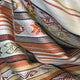 Fabric with horizontal stripes and intricate geometric patterns in shades of orange, red, black, green, and cream, folded and layered closely, creating a textured appearance.