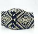 A fabric face mask displays a bold black and beige geometric pattern, featuring diamond shapes and angular lines, set against a plain white background.