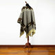 A poncho is draped over a wooden mannequin stand. It features brown and beige geometric patterns, fringed edges, and small floral motifs, set against a plain white background with wooden flooring.