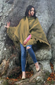 A person wearing an olive-green shawl and blue jeans stands against a large, textured tree trunk, surrounded by fallen leaves, creating an autumnal atmosphere.