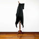 A black fringed cloak with a hood is draped over a wooden mannequin stand, set against a white wall and a hardwood floor.