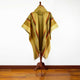 A woven poncho with a hood, featuring geometric patterns and earthy tones, is displayed on a wooden stand in a room with hardwood flooring and a plain white wall.