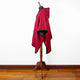 A red cape with black trim drapes on a wooden mannequin stand, set against a white wall with a wooden floor.