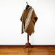 A brown poncho adorned with white geometric patterns drapes over a mannequin, standing on a wooden floor against a plain white wall.