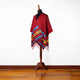 A red poncho with colorful patterns and fringes is displayed on a wooden mannequin stand in a room with wooden flooring and a plain white wall.