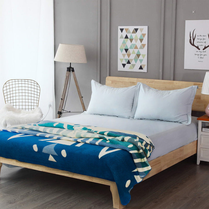 Bed with blue and teal geometric blanket rests in a modern bedroom. A lamp, wire chair, and artwork featuring triangles and antlers decorate the gray-walled space. Artwork text: to the moon and back.
