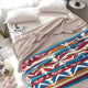 Bed with white pillows and a beige blanket partially covering a colorful geometric-patterned throw, in a sunlit bedroom. Nearby, a small table holds a magazine and plant, with slippers beside.
