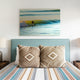 Nongora baby alpaca wool blanket – two patterned cushions rest on a bed with a striped blanket, in a bedroom setting. Above, a wall-mounted photograph shows a surfer riding a wave at sunset