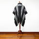 Mannequin displays black-and-white striped poncho with a hood, resting on a wooden stand against a white wall and wooden floor.