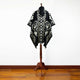 A mannequin displays a black and white poncho with intricate geometric and eagle designs, standing indoors on a wooden floor against a plain white wall.