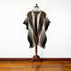 A woolen poncho with geometric patterns in brown and cream hangs on a wooden stand, set against a plain white wall and wooden floor.