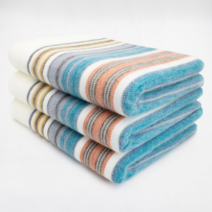 Nongora baby alpaca wool blanket – three folded blanket display colorful horizontal stripes in blue, orange, yellow, and gray hues resting on a clean, white surface, suggesting a neat arrangement