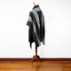 A black and gray striped poncho drapes over a wooden mannequin stand, set against a white wall and hardwood floor.