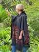 A person in a patterned black poncho touches large green leaves, surrounded by lush tropical plants and vibrant flowers.