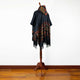 A dark poncho with colorful geometric patterns hangs on a wooden stand, displayed against a plain white wall on hardwood flooring.