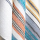 Nongora baby alpaca wool blanket – a close-up of a woolen fabric featuring diagonal stripes in orange, blue, and gray hues. The fabric is draped, emphasizing its texture and color pattern in a soft light