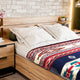 Bed with white pillows and a colorful patterned blanket, in a room with a light wood headboard and side tables. Brick wall and soft lighting provide a cozy setting.