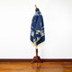 A navy blue patterned poncho drapes over a wooden mannequin stand. The poncho features white geometric designs and fringe, set against a white wall and wooden floor.