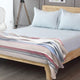 Pindo baby alpaca wool blanket – a neatly made bed with light blue sheets and pillows. A striped blanket lies on top. The setting is a modern bedroom with a wooden floor and a tripod lamp