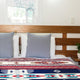Bed with two gray pillows resting against a wooden headboard, featuring a colorful geometric-patterned blanket. A small potted plant sits on a bedside table in a bright room.