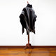 A dark poncho with gray stripes drapes over a wooden mannequin stand, set against a white wall and wooden floor.