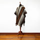 A woven poncho with geometric patterns drapes over a mannequin stand, displayed in a room with wooden flooring and a plain white wall.