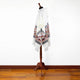 A white poncho with colorful geometric patterns drapes over a wooden mannequin stand, set against a plain white wall and hardwood floor.