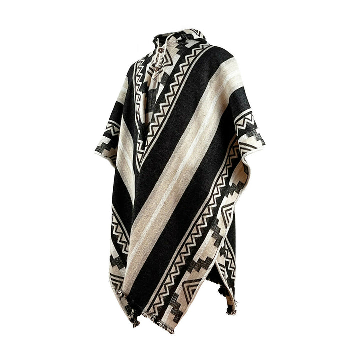 A wool poncho with black and cream zigzag patterns and broad stripes, hanging against a white background, features a fringed hem and a round neck opening.