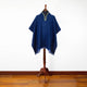 A blue poncho with a patterned collar rests on a wooden stand. The setting features a wooden floor and a plain white background.