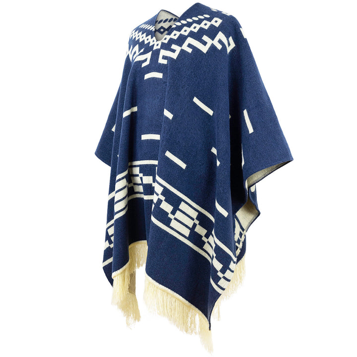 A blue poncho with white geometric patterns hangs in a studio setting. The poncho features fringe detailing at the bottom and a V-neck opening.