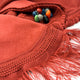 A red textured fabric with fringe lies partially open, revealing colorful small pebbles inside. The fabric features intricate stitching and a smooth, woven texture on a flat surface.