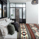 Bed adorned with patterned blanket and multiple pillows, situated in a modern bedroom with a large window. In the background, a glass-walled shower enclosure is visible.