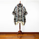 A mannequin displays a hooded poncho with intricate geometric patterns in brown, gray, and cream, hanging in a room with a wooden floor and white wall background.