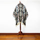 A patterned poncho with a hood hangs on a wooden mannequin stand, displayed against a plain white wall on a polished wooden floor.