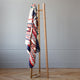 A wooden coat stand holds a colorful, patterned blanket draped over it, in a minimalist room with a light gray wall and herringbone wood floor.