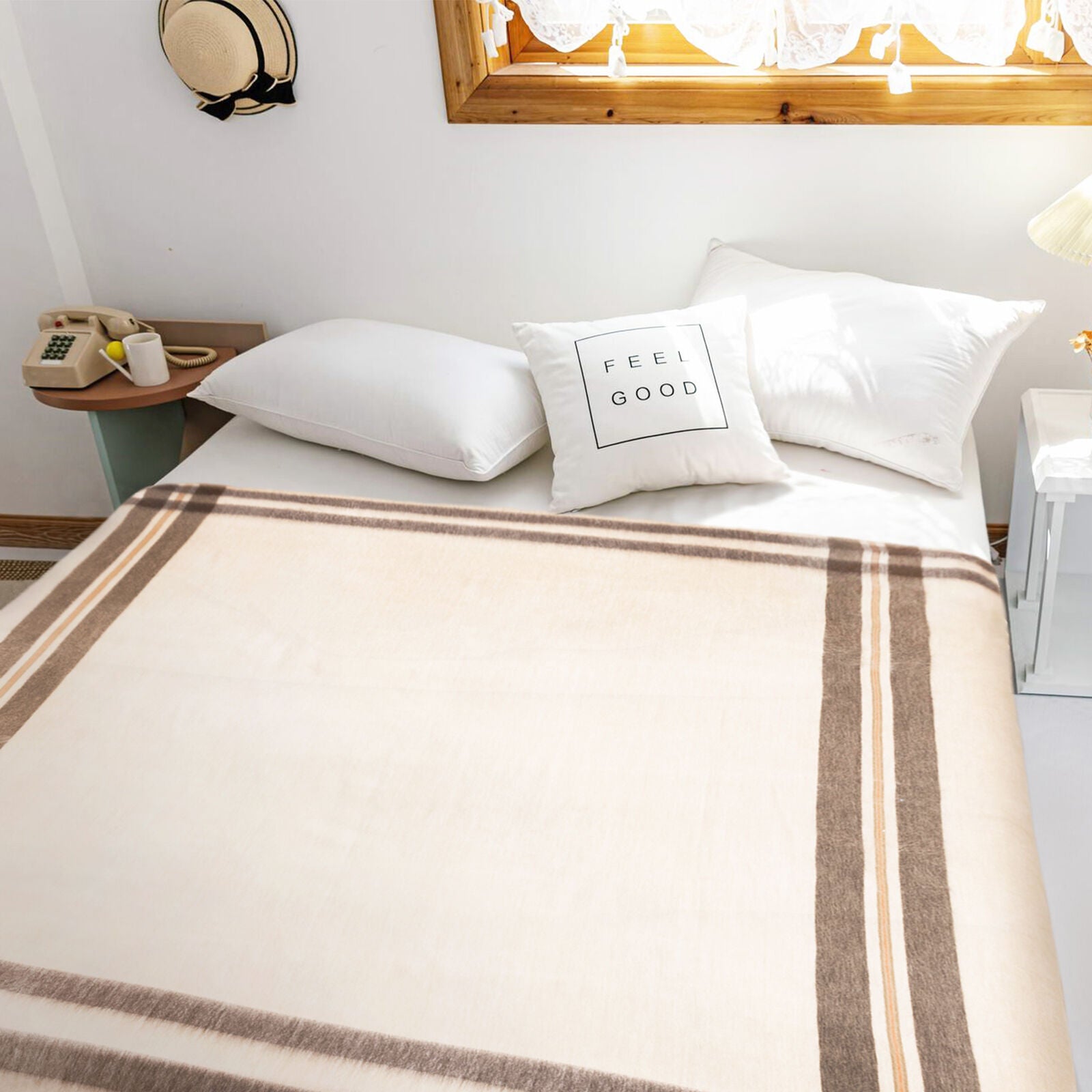 Bed with a neatly arranged cream and brown-striped blanket, white pillows including one with FEEL GOOD text, in a cozy room with a wooden window, hat, phone, and nightstand.